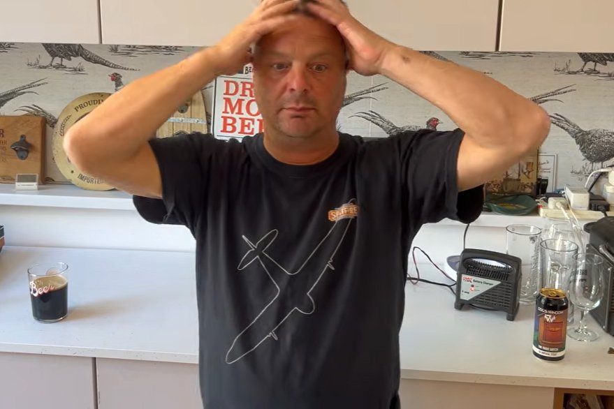 A man wearing a black T-shirt with a Spitfire outline stands behind a counter with both hands on his head, looking directly at the camera with a surprised expression.